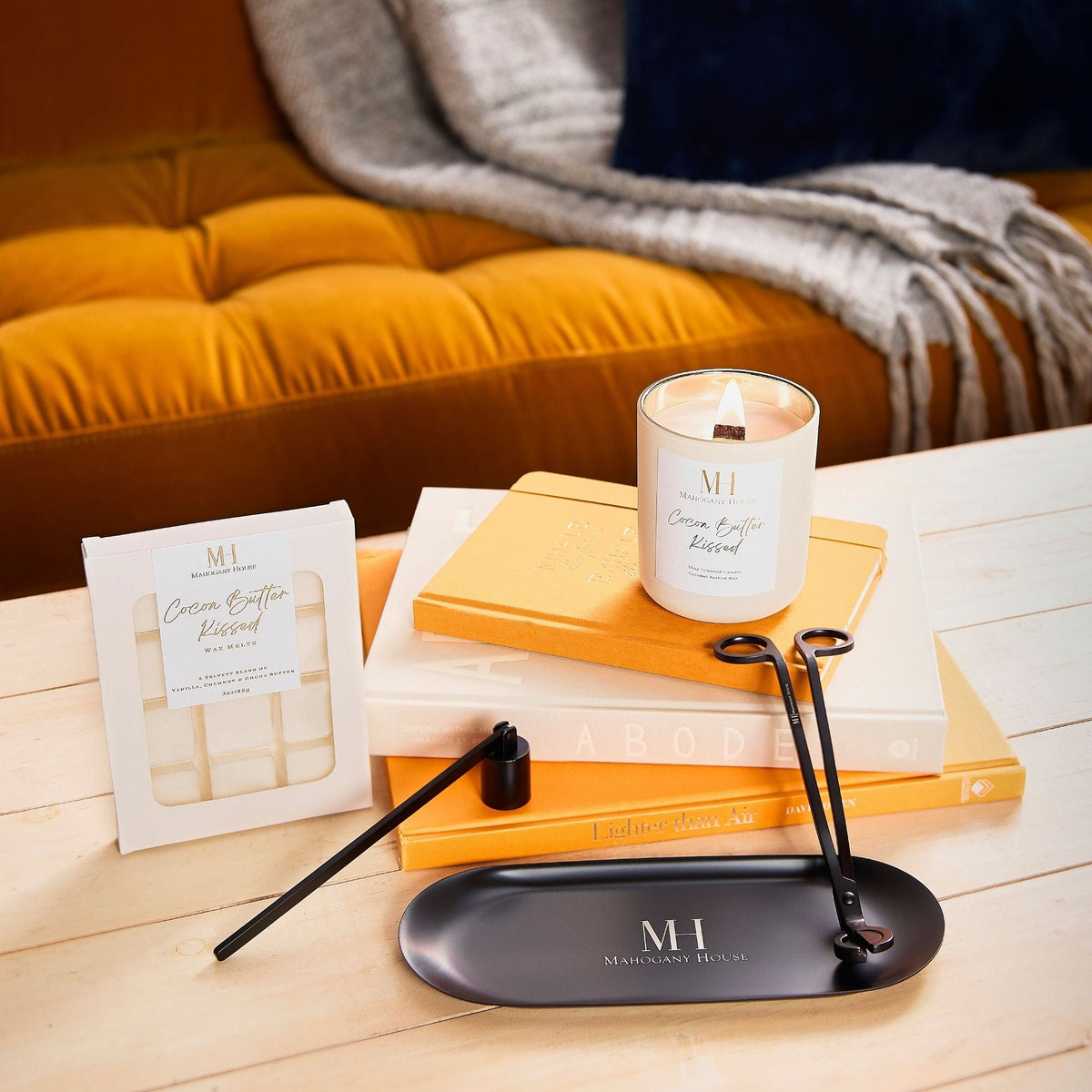 Lit Cocoa Butter Kissed candle on stacked books beside wax melts and black candle-care tools on a coffee table.